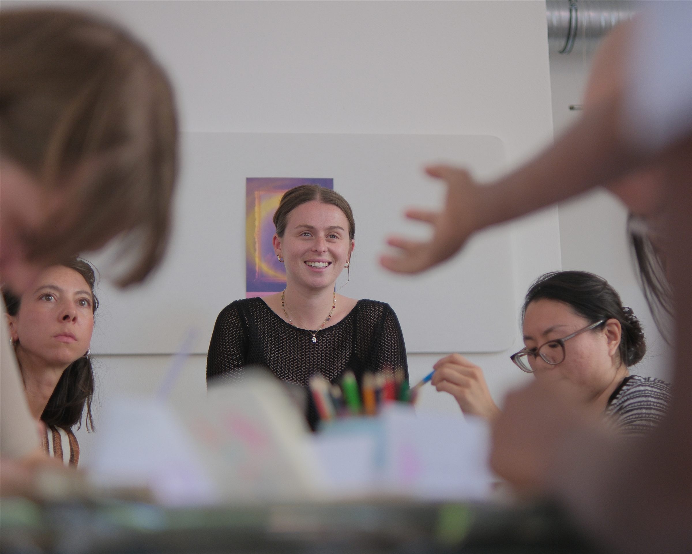 Smiling workshop leader seated at a table with participants, a cup of colored pencils in the foreground and blurred hands and heads framing the shot